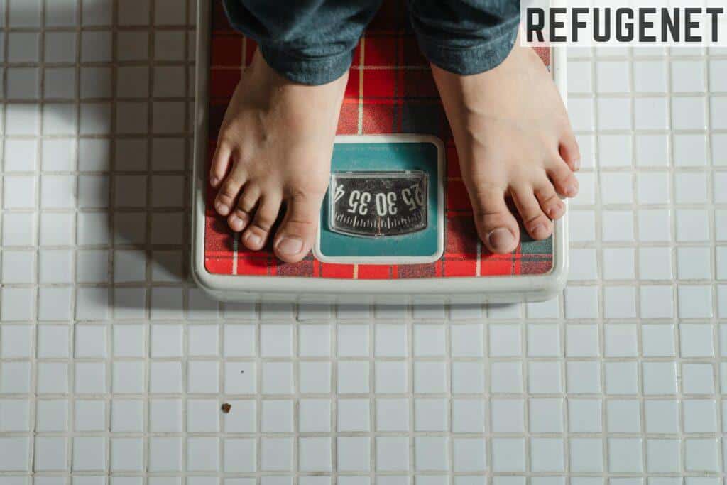 From above crop anonymous barefoot child in jeans standing on weigh scales on tiled floor of bathroom