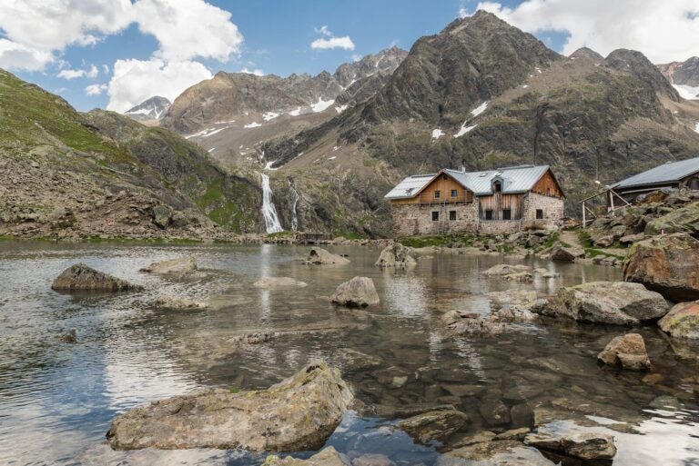 Refugio de montaña junto a un lago de alta montaña, con picos nevados al fondo